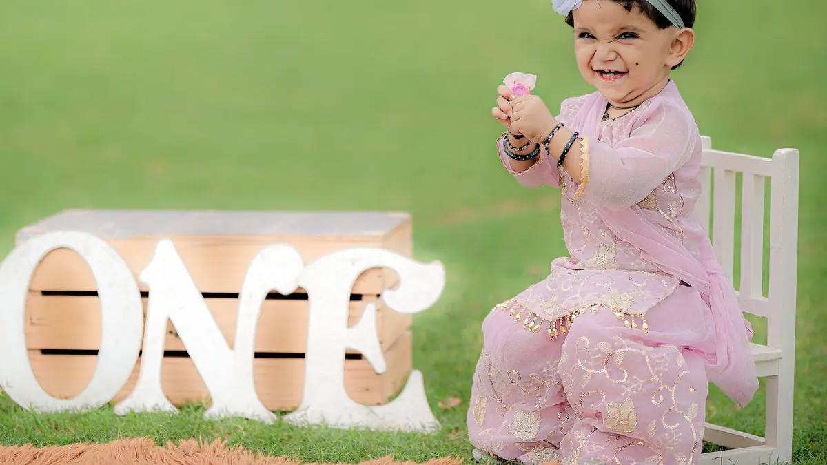 A baby girl in a pink outfit sitting beside large ONE letters on a lawn
